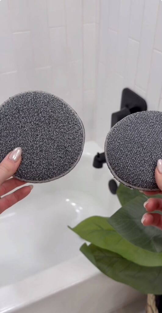 Two hands hold up round, gray textured scrub pads over a white bathtub with a black faucet. A green plant is visible in the bottom right corner of the image.