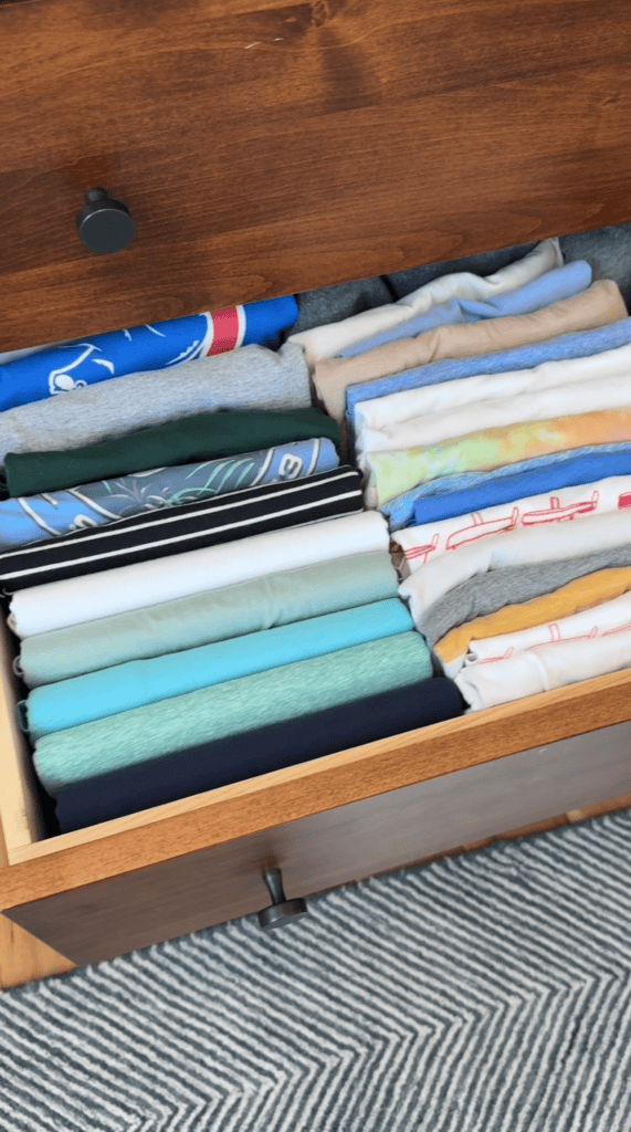 An open wooden drawer neatly filled with folded t-shirts arranged upright in rows, showing different colors and patterns. A chevron-patterned rug is partially visible on the floor below.