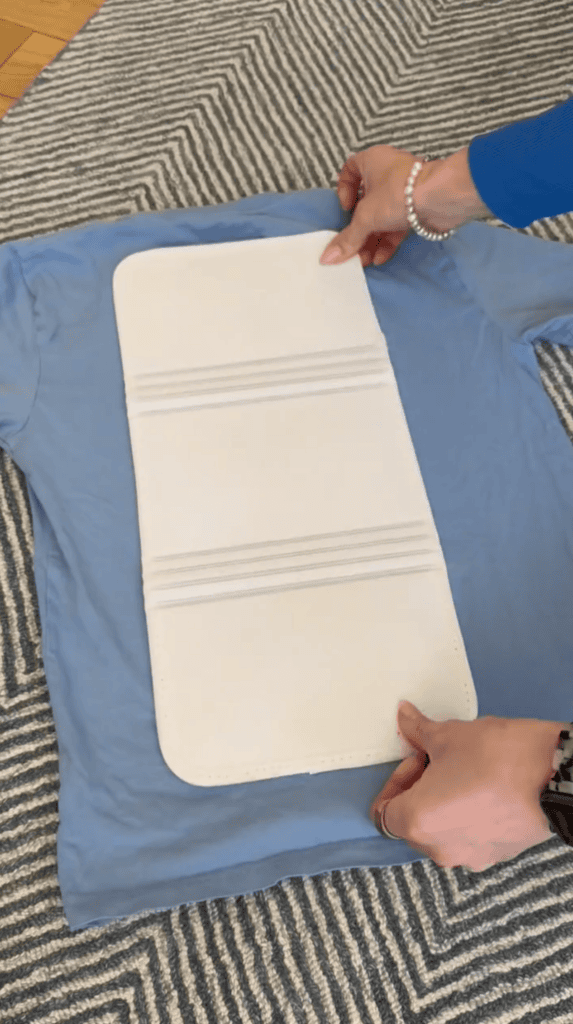 A person places a folding board on a light blue T-shirt, which is laid flat on a gray and white patterned rug.