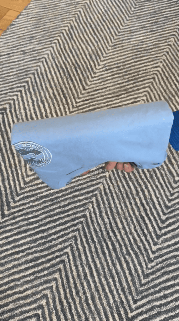 A hand is holding a rolled-up light blue T-shirt with a circular white logo, against a patterned black, white, and gray rug background.