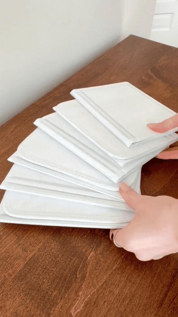 A person arranges a stack of white, rectangular cloth napkins or cleaning cloths on a brown wooden table near a light-colored wall.