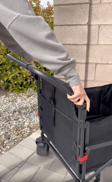A person wearing a gray long-sleeve shirt is folding or adjusting the handle of a black collapsible utility wagon on a paved surface near a stone wall and some greenery.