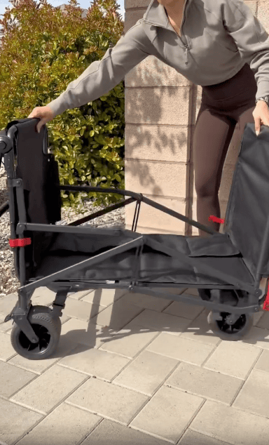 A person wearing a gray zip-up jacket and brown leggings is unfolding a black collapsible wagon on a paved outdoor area, with greenery and a brick wall in the background.