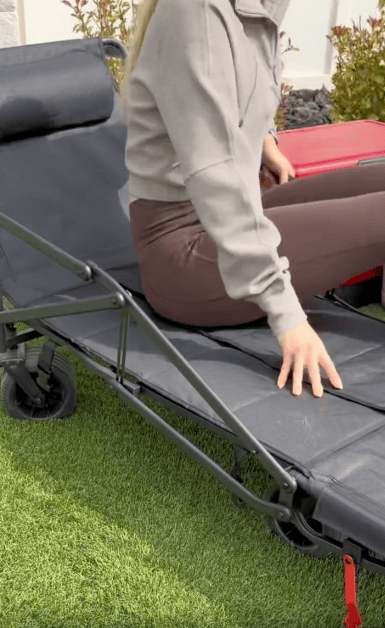 A person in a gray hoodie and brown leggings sits on a black foldable lounge chair with wheels, placed on grass. Shrubs and a white wall are visible in the background.