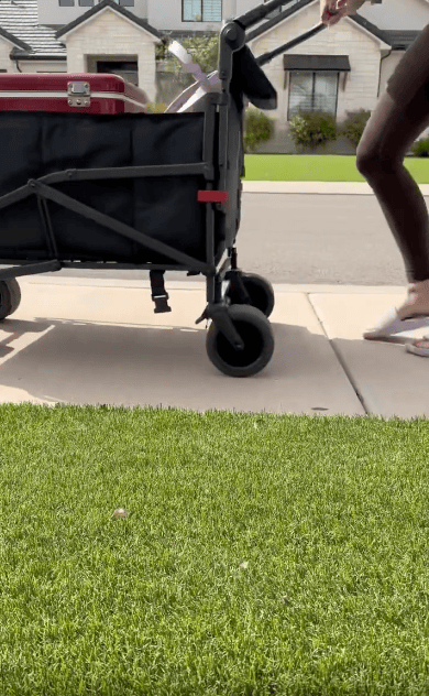 A person wearing sandals pushes a black collapsible wagon with a red suitcase inside along a sidewalk next to a lawn, with houses visible in the background.