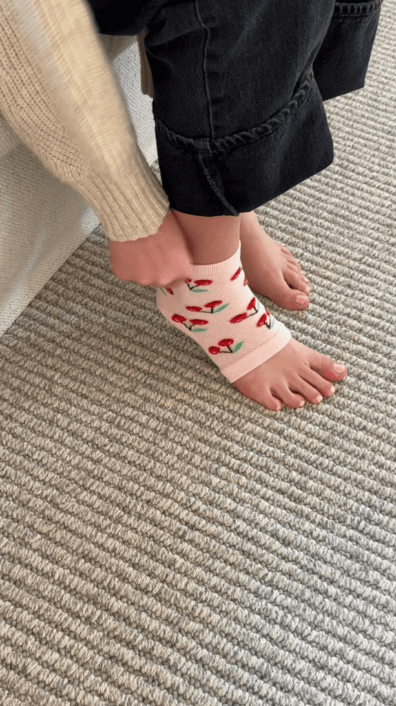A person sitting on a light-colored sofa adjusts a pale pink sock with red cherries on their foot. Their other foot is bare, and they are on a textured, light-colored carpet.