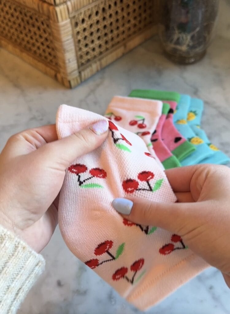 A person with light purple nail polish holds a pink sock with a red cherry pattern. Other colorful socks with fruit designs are stacked on a marble countertop in the background.
