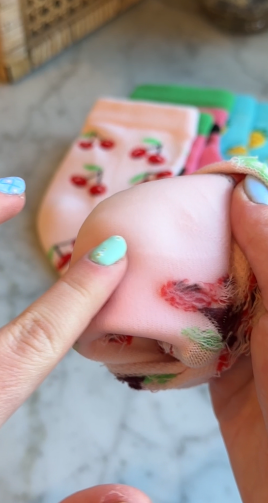 A close-up of hands with blue painted nails holding a pink sock with a cherry pattern, showing a hole in the fabric while other colorful socks are visible in the background.