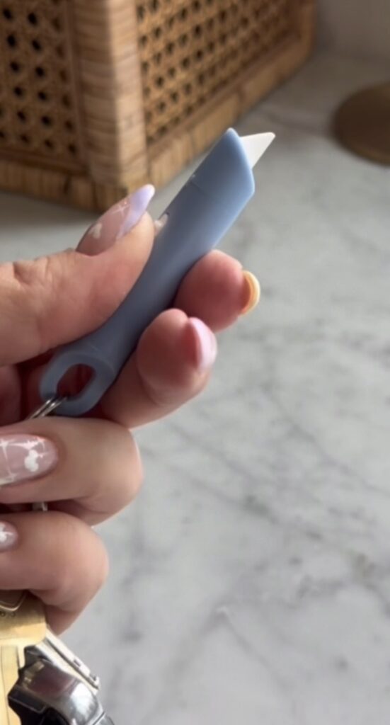 A hand with pastel-colored, decorated nails holds a small blue tool with a white tip and a key ring hole, against a blurred marble countertop background.