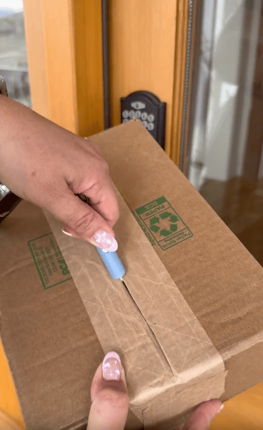 Person opening a cardboard package with a small blue tool in front of a wooden door with a keypad lock. The box has recycling symbols printed on it.