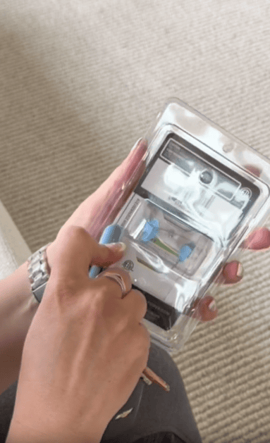 A person’s hands holding a packaged medical device with blue and white components, appearing to open it with a small tool. The background is a textured beige carpet.