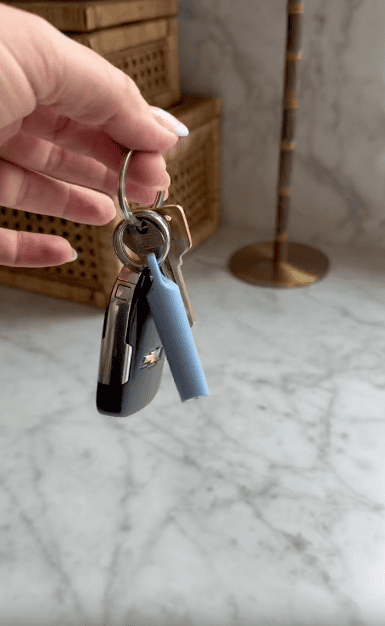 A hand holds a keyring with a car key, a house key, and a blue cylindrical object against a marble surface and rattan boxes in the background.