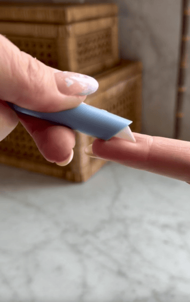 A hand holds a blue silicone nail tool, gently pushing back the cuticle on another person's fingernail over a light marble surface.