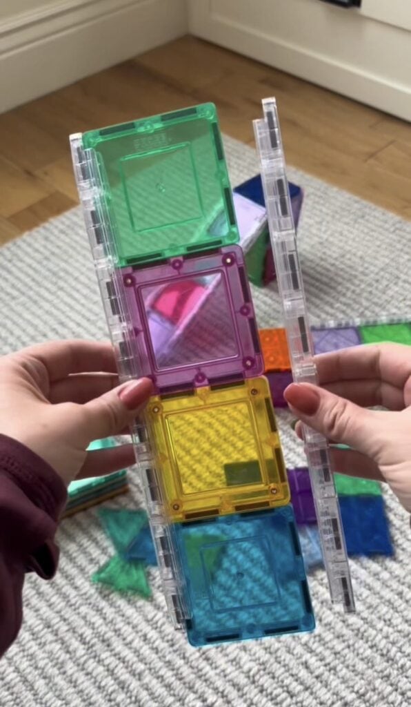 Hands hold a structure made of colorful magnetic tiles, featuring green, purple, yellow, and blue squares. More loose tiles are scattered on a gray carpet and wooden floor in the background.