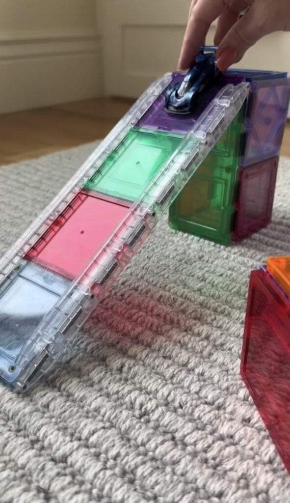 A hand pushes a small toy car up a ramp made of colorful translucent magnetic tiles on a textured carpet.