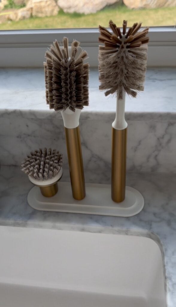 Three gold and white kitchen brushes of varying sizes are standing upright in a white holder on a marble countertop near a sink.