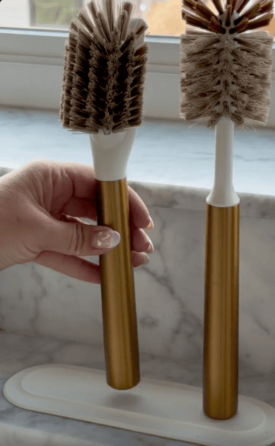 A hand holds a gold-handled bottle brush with brown bristles next to a similar brush on a white holder, set against a marble countertop and window background.