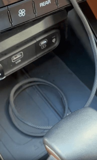 A close-up of a car’s center console area, showing USB ports, a 12V outlet, and a round black charging cable resting in the compartment below. Part of the gear shifter and climate control buttons are visible.