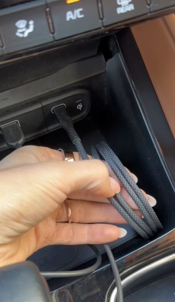 A hand holding a braided charging cable near a car's dashboard, with the cable plugged into a USB port below the air conditioning controls.