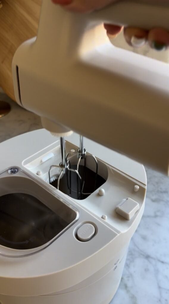 A close-up of a white electric hand mixer with metal beaters inserted into a matching white mixing bowl on a marble countertop. A person's hand is holding the mixer.