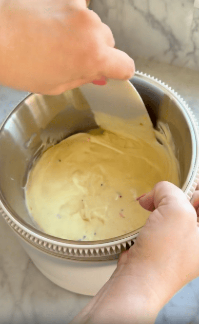 A person mixes cake batter with rainbow sprinkles in a metal bowl using a spatula, with both hands visible against a marble countertop background.