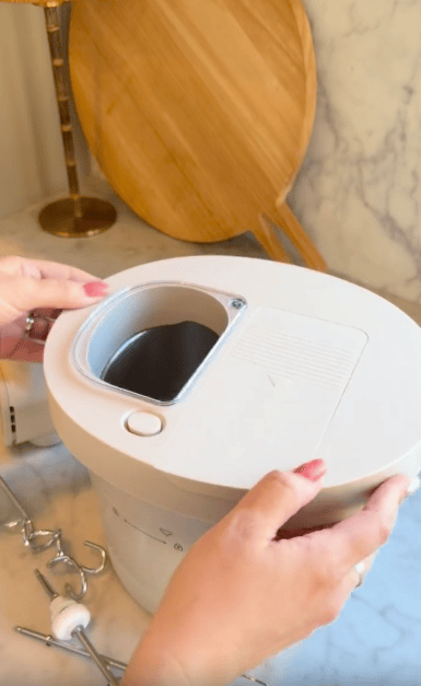 A person with pink nails holds the lid of a kitchen appliance with a windowed opening, placed on a marble countertop next to metal mixer attachments and a round wooden cutting board.