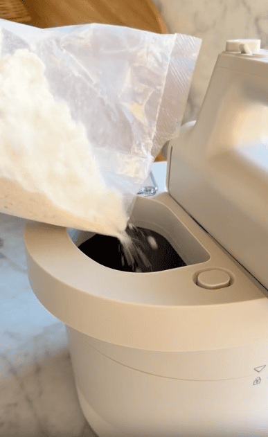 A hand pours flour from a clear plastic bag into the mixing bowl of a stand mixer on a marble countertop.