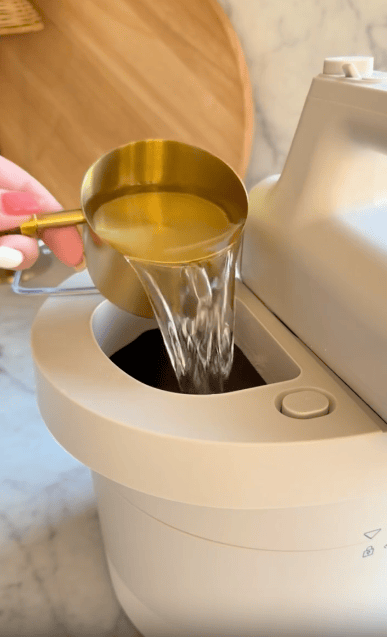 A hand pours water from a gold measuring cup into the open top of a white kitchen appliance, possibly a bread maker, on a marble countertop.
