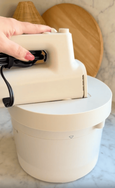 A hand places a white electric hand mixer with a coiled black cord onto a matching white appliance base, with wooden kitchen boards in the background.