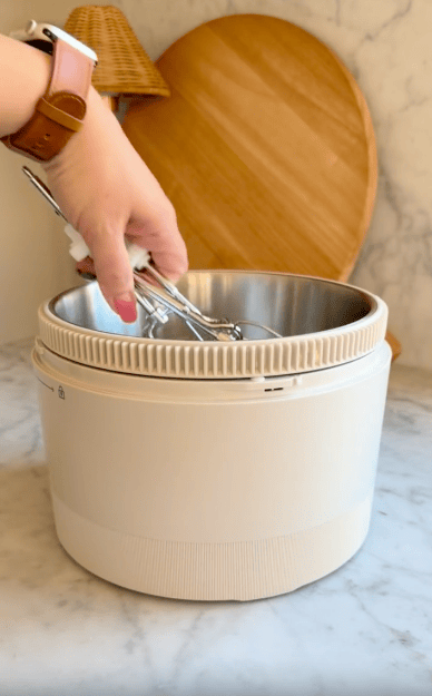 A hand holding tongs hovers above a beige and stainless steel appliance on a marble countertop, with wooden kitchen decor in the background.
