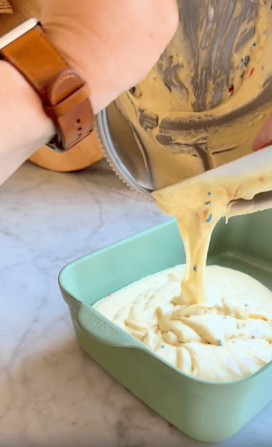 A hand pours cake batter with colorful sprinkles from a mixing bowl into a square green baking dish on a marble countertop.