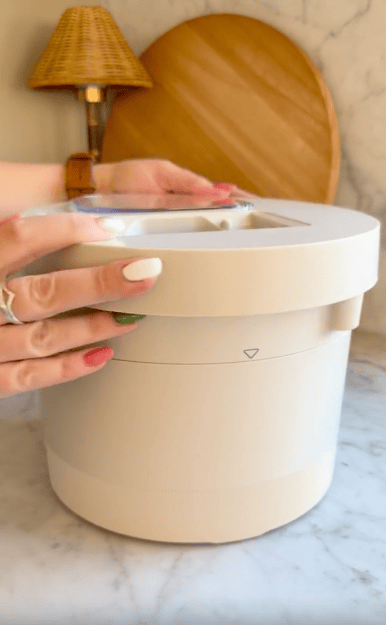 A person with painted nails is placing the lid on a round, beige kitchen appliance on a marble countertop, with wooden decor and a lamp in the background.