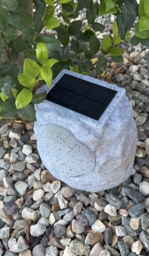 A solar-powered outdoor speaker shaped like a gray rock sits on gravel near green leafy plants. The speaker has a solar panel on top and a mesh speaker grill on the side.