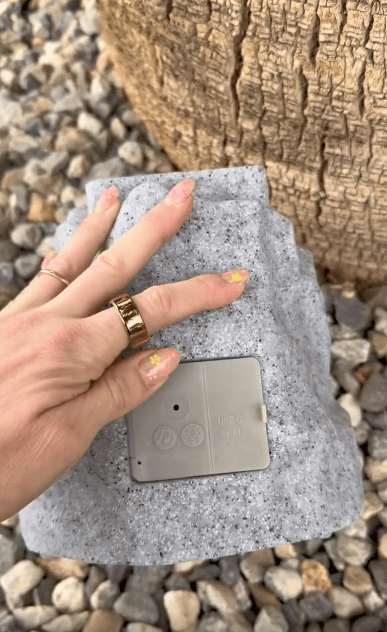 A hand with painted nails lifts a fake rock covering an electrical outlet with USB ports, set among small stones at the base of a tree.