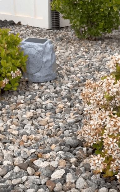 A garden area with small, multicolored rocks, flowering bushes, and a gray, rock-shaped solar light. A white fence and green shrubbery are in the background.