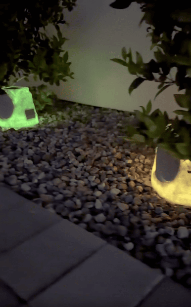 Two small garden lights, one glowing green and one warm yellow, illuminate bushes planted in a bed of rocks beside a paved surface at night.