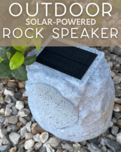 A solar-powered outdoor speaker designed to look like a gray rock sits among small stones and leaves, blending into a garden setting. Text above reads, "Outdoor Solar-Powered Rock Speaker.