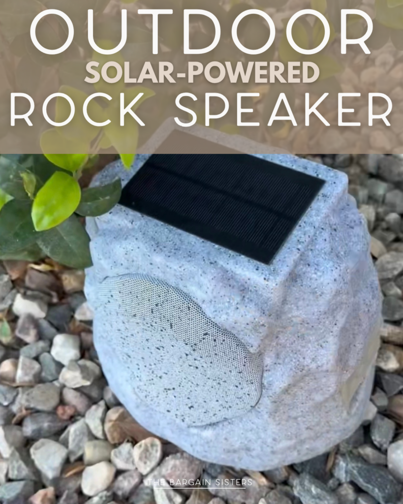A solar-powered outdoor speaker designed to look like a gray rock sits among small stones and leaves, blending into a garden setting. Text above reads, "Outdoor Solar-Powered Rock Speaker.