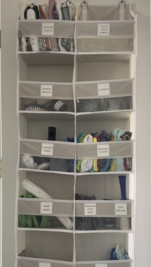 Hanging organizer with labeled shelves containing items such as towels, shower brushes, candles, steam mop parts, washing cloths, mop pads, stain remover, and a lint roller.