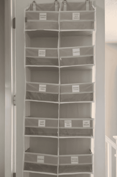 A white hanging organizer with clear pockets attached to a door, each pocket labeled for items like towels, sponges, mop pads, steam mop pads, and cleaning supplies in a well-lit hallway.