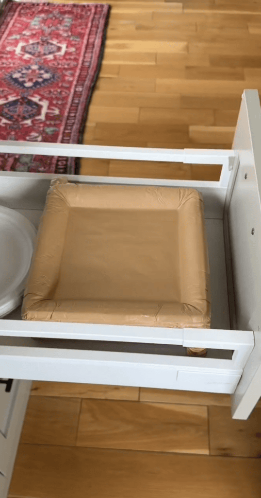 A drawer is open, showing a stack of square, tan-colored paper plates and a stack of round white plates inside a white cabinet. A wooden floor and a patterned red rug are visible in the background.