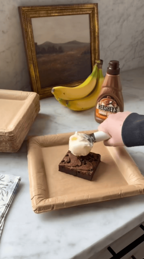 A hand scoops vanilla ice cream onto a brownie on a square paper plate. In the background are bananas, a bottle of Hershey’s chocolate syrup, a framed landscape painting, and more paper plates.