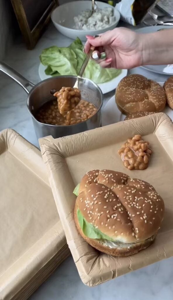 A hand spoons baked beans from a pot onto a square paper plate next to a sandwich with lettuce in a sesame seed bun. Other buns and lettuce are visible in the background.
