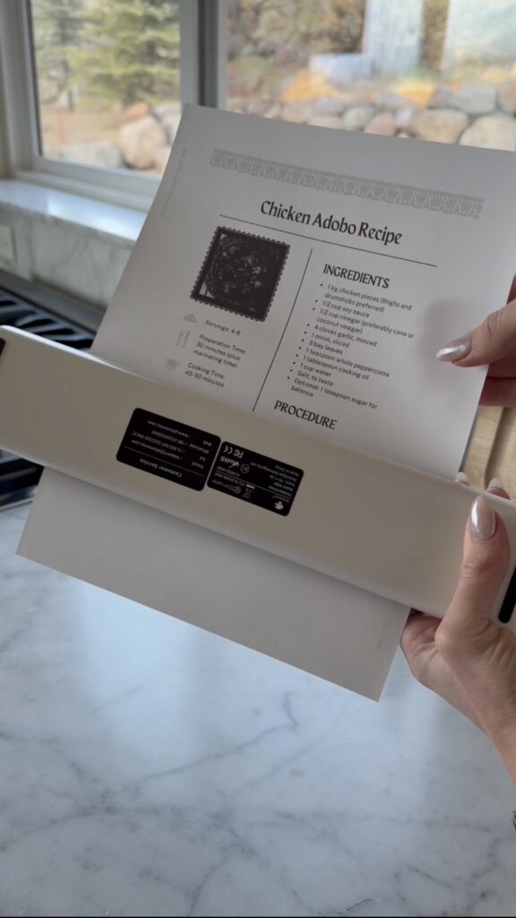 A person holds a sheet of paper with a Chicken Adobo recipe, placing it into a white laminating machine on a marble countertop. The recipe lists ingredients and a small photo of the finished dish.