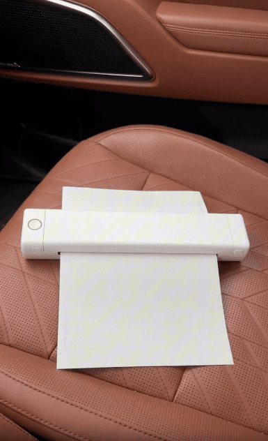 A portable document scanner sits on brown perforated leather car seat, scanning a sheet of white paper.