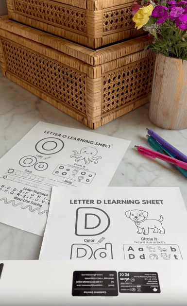 Two letter learning sheets on a marble table: one for letter O with an octopus, one for letter D with a dog. Colorful pens, wicker baskets, and a vase of flowers are also visible.