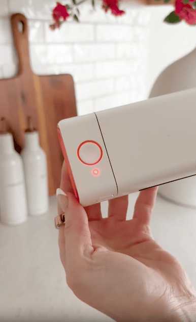 A hand holds a white electronic device with a red power button and indicator light illuminated. In the background, there are two white bottles, a wooden cutting board, and a white vase on a kitchen counter.