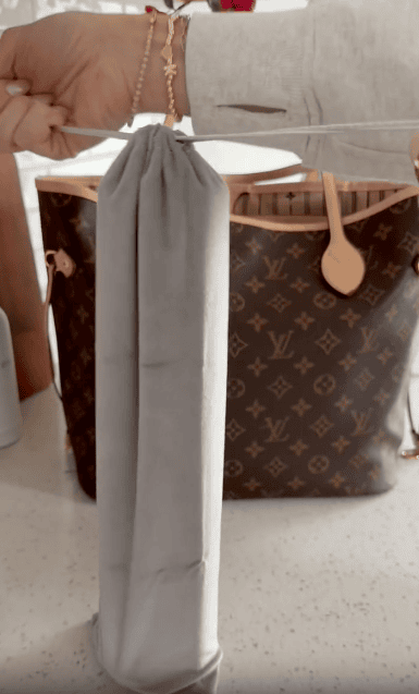 A hand holds a grey drawstring pouch by the strings, with a brown Louis Vuitton monogram tote bag in the background on a white surface.