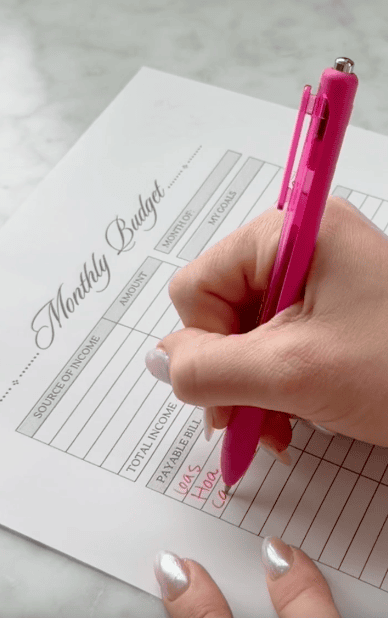 A person writes “gas,” “hoa,” and “car” in pink ink on a monthly budget sheet with columns for income and expenses, using a pink pen on a marble surface.