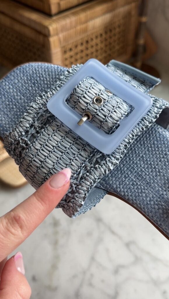 A close-up of a hand with light pink nail polish pointing at a textured, blue woven sandal with a large, square blue buckle.